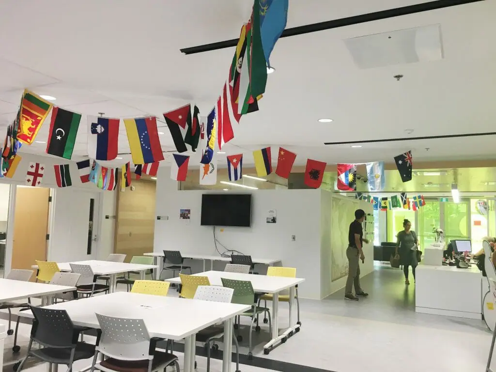 In this image: inside the Dalhousie University International Centre. Different countries' flags hang from the ceiling. A cluster of empty tables and chairs.