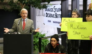 In this image: MacKinnon speaks while the student protesters silently hold their signs.