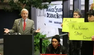 In this image: MacKinnon speaks while the student protesters silently hold their signs.