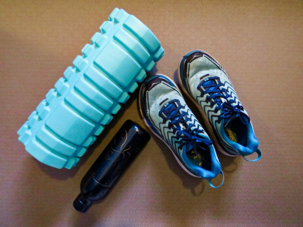 In this image: A foam roller, a water bottle and a pair of running shoes.