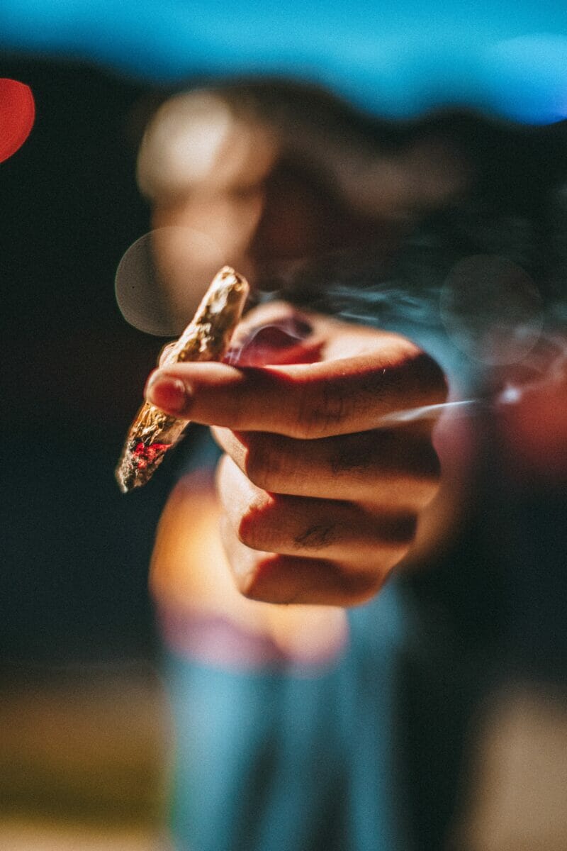 In this image: a blurred out person holds a lit joint up.