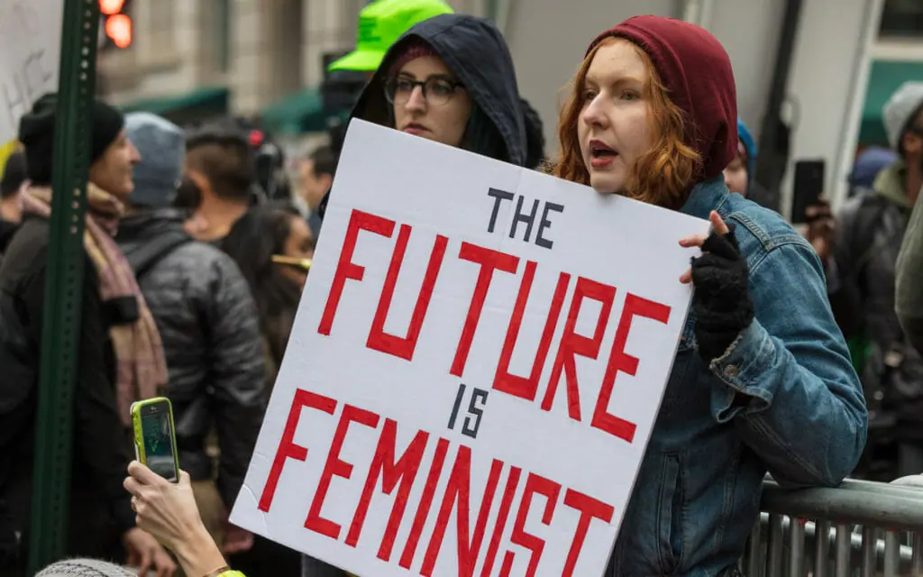 In this image: A woman holds a sign at a rally that says "the future is feminist."
