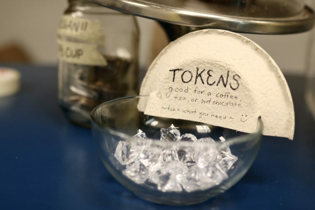 In this image: A glass jar filled with gems with a sign saying "Tokens / good for a coffee, tea, or hot chocolate / ~take what you need~