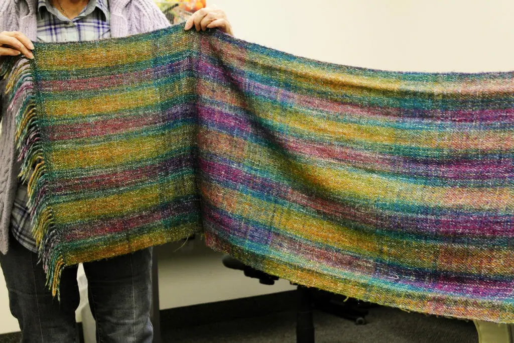 In this image: A person holds up a hand-woven scarf made by Helene Comstock.