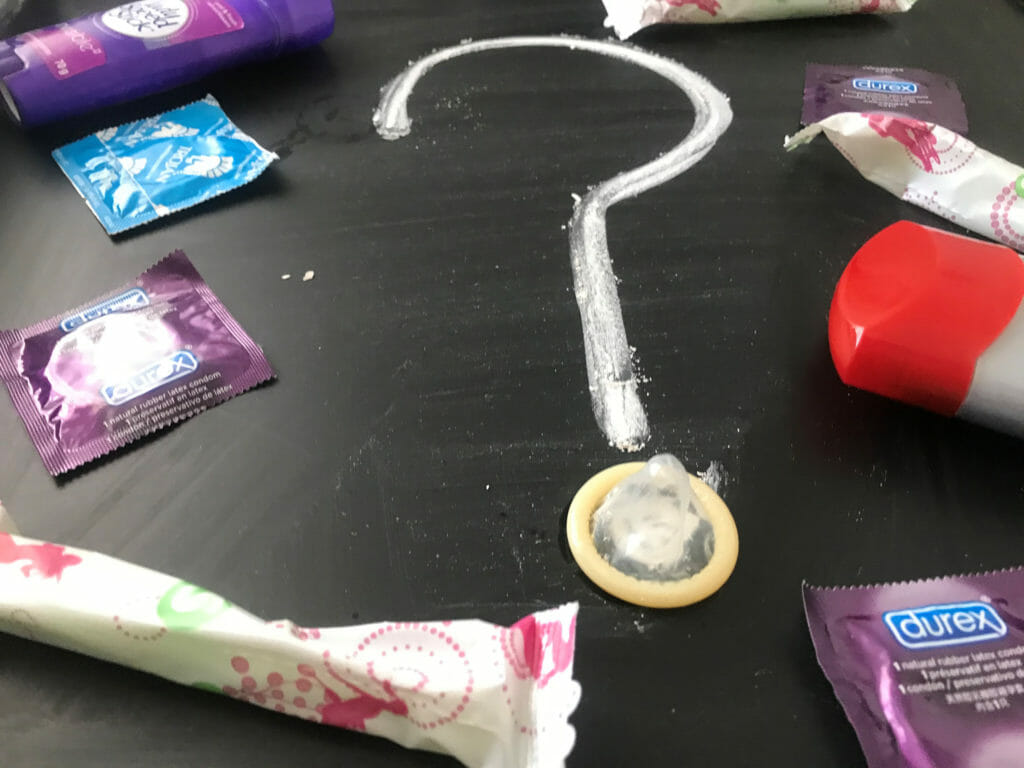 In this image: Condoms, tampons and a deodorant stick surround a question mark made with chalk.