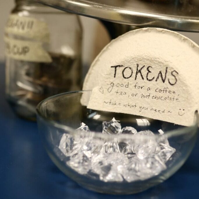 Galleytokenprogram_ChrisStoodley In this image: A glass jar filled with gems with a sign saying "Tokens / good for a coffee, tea, or hot chocolate / ~take what you need~
