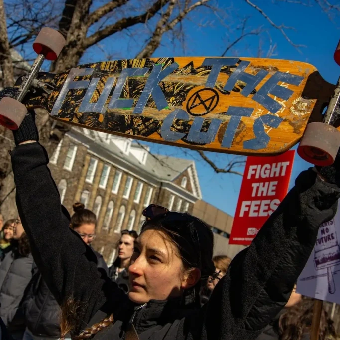 An individual raises a longboard with a message on it made of tape outside the Henry Hicks Academic Administration Building on Dalhousie University campus in Halifax, Nova Scotia on March 18, 2026. (Lukas Kohler/The Dalhousie Gazette)