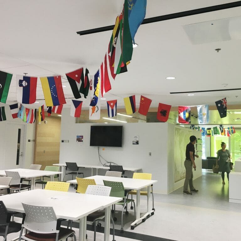 In this image: inside the Dalhousie University International Centre. Different countries' flags hang from the ceiling. A cluster of empty tables and chairs.