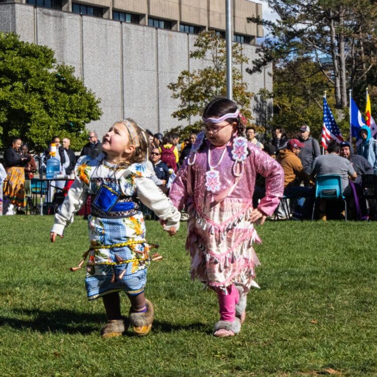 Two Mi’kmaw children holding hands while dancing in their regalia at Dalhousie’s 14th annual Mawio’mi on the Dal quad in Halifax, Nova Scotia on October 16, 2024. The girls show their cultural upbringing as each piece of regalia is typically handcrafted and holds deep cultural and spiritual significance for the wearer and their community (Image by Ainslie Nicholl-Penman)