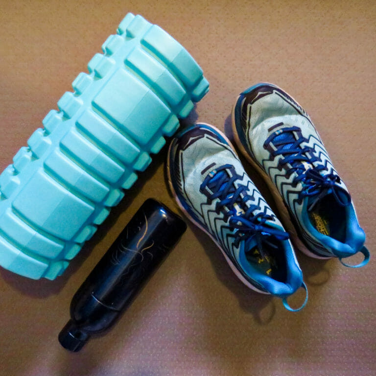 In this image: A foam roller, a water bottle and a pair of running shoes.