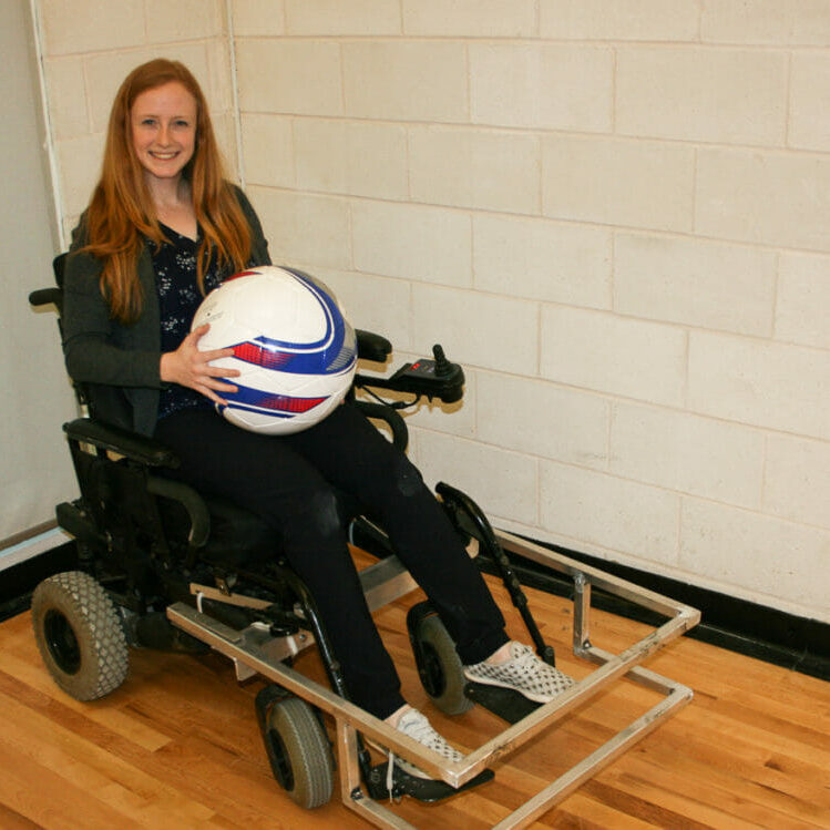 In this Image: Madison Campbell sits in a power wheelchair holding a powerchair soccer ball.