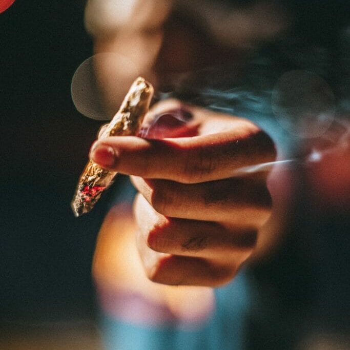 In this image: a blurred out person holds a lit joint up.